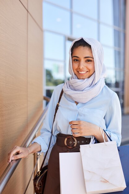 vertical portrait of beautiful middle-eastern woman holding shopping bags and smiling at camera while leaving mall with purchases