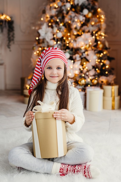 Vertical portrait of beautiful pleasant looking small child wears knitted sweater and socks, sits crossed legs with present, has desire to wrap it, being in living room near decorated new year tree