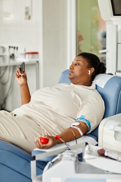 Vertical portrait of black young woman giving blood while laying in chair at blood donation center a
