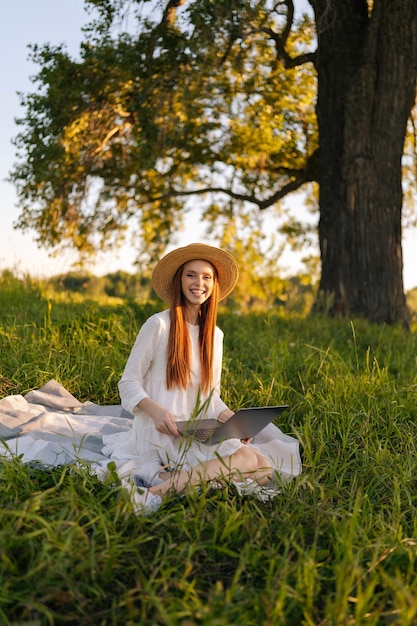 Vertical portrait of charming smiling young female freelancer in straw hat and white dress sitting with laptop looking at camera on field of green grass