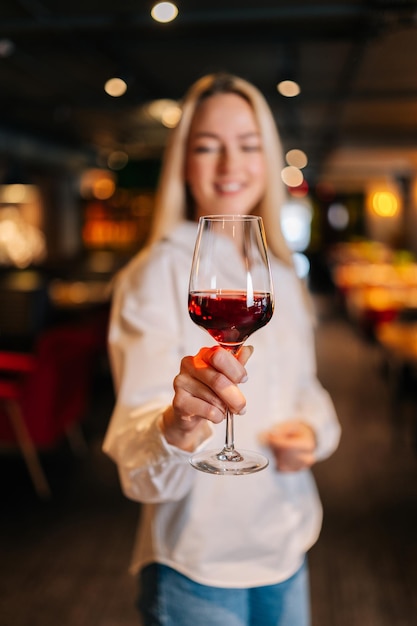 Vertical portrait of cheerful blonde woman holding glass of red wine standing in restaurant with luxury interior looking away