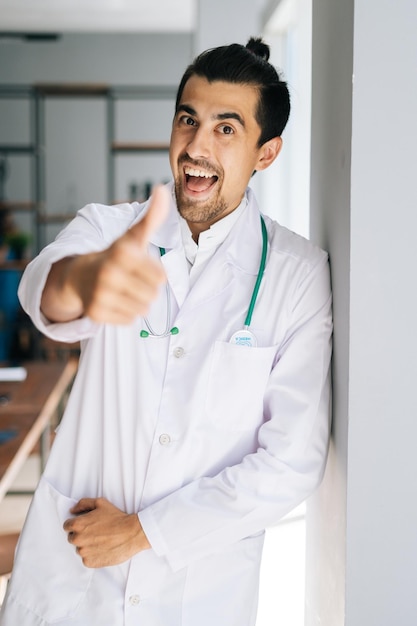 Vertical portrait of cheerful male doctor in white medical uniform showing thumbup gesture in hospital office looking at camera