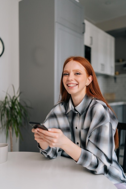 Vertical portrait of cheerful young woman sitting at table sharing good news at social media via smartphone smiling looking at camera Happy female reading text