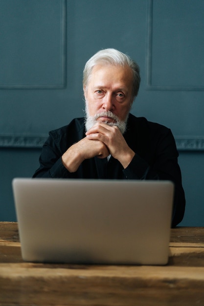 Vertical portrait of confident bearded mature business man sitting at wooden table with laptop computer at home office looking at camera