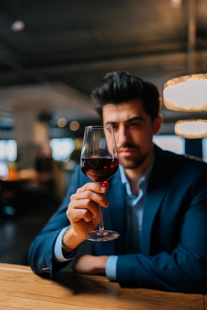 Vertical portrait of confident elegant man in fashion suit holding glasses of red wine sitting at table in restaurant with dark interior looking at camera