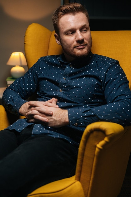 Вертикальный портрет уверенного в себе красивого мужчины в стильной рубашке, удобно сидящего в роскошном желтом кресле со скрещенными руками в темной уютной гостиной с современным интерьером, смотрящим в камеру