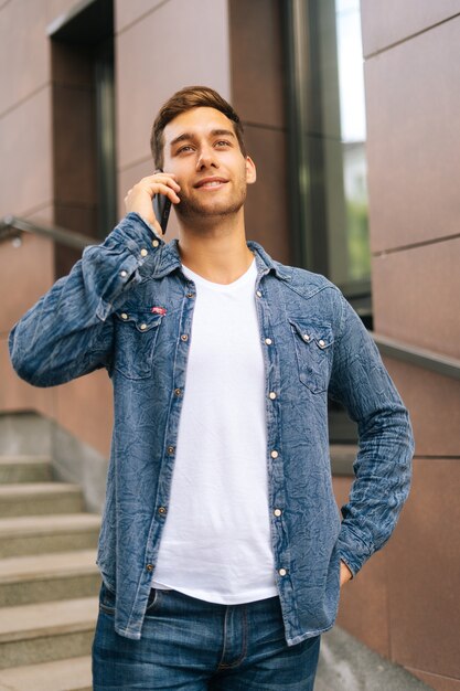 Vertical portrait of confident handsome young man talking on mobile phone standing on stairs of modern office building, blurred background. Successful businessman speaking on smartphone outdoors.