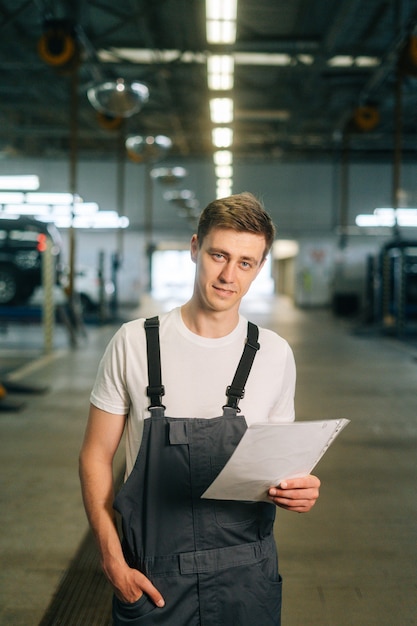 Vertical portrait of confident handsome young mechanic male wearing uniform holding clipboard