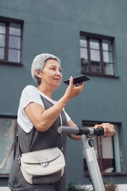 Vertical portrait of contemporary mature woman speaking by smartphone while riding electric scooter in city