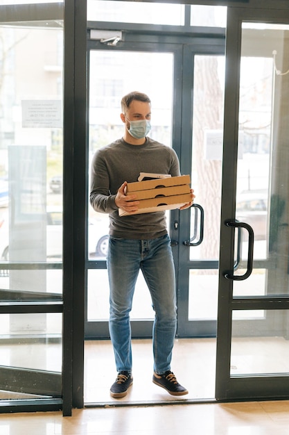 Vertical portrait of courier male in medical face mask holding\
boxes hot pizza and payment pos terminal entering lobby of\
apartment building