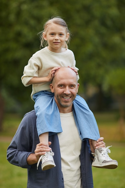 Vertical Portrait of cute girl sitting on dads shoulders and enjoying walk in park