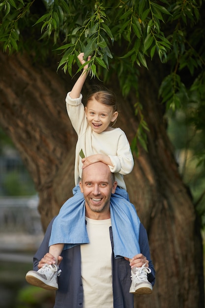 Vertical Portrait of cute little girl laughing happily while sitting on dads shoulders and enjoying walk in park