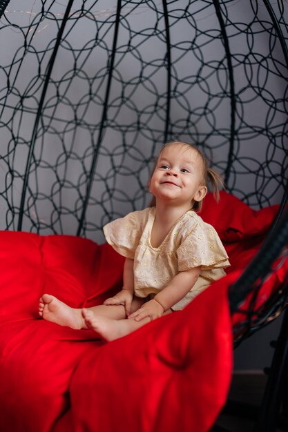 vertical portrait of cute toddler girl relaxing in redcushioned hanging chair gesturing playfully
