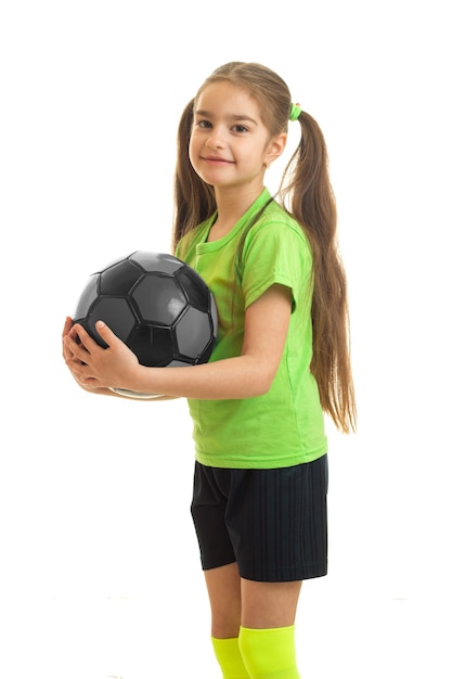 白い背景で隔離の手でサッカーボールを保持している緑のシャツのかわいい女の子の縦の肖像画