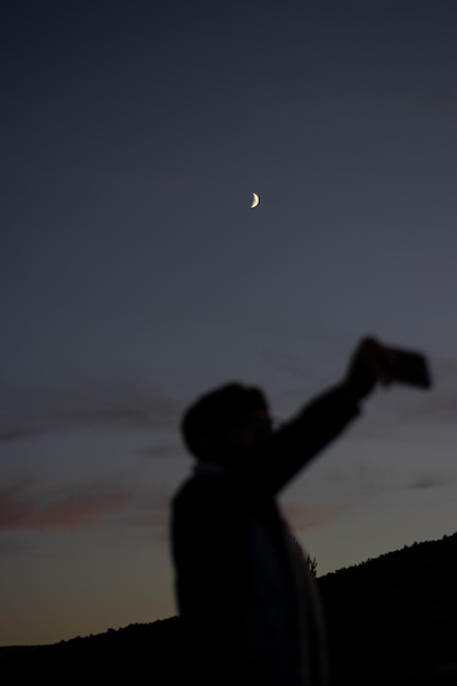 Vertical portrait of the defocused silhouette of a man taking a selfie with the moon at dusk focus on the moon