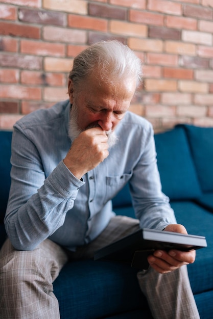 Vertical portrait of depressed elderly bearded man sitting on sofa at home looking at picture of wife in frame and crying