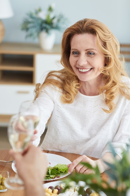 Vertical portrait of elegant mature woman raising champagne glass and toasting while enjoying romantic dinner