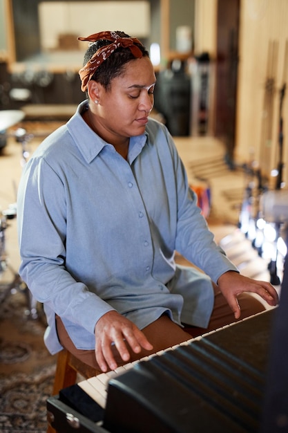 Vertical portrait of ethnic young woman playing keyboard while composing music in recording studio