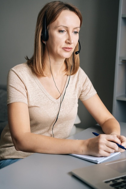 vertical portrait of focused female operator talking with headsets and consulting client at home office sitting at table with laptop