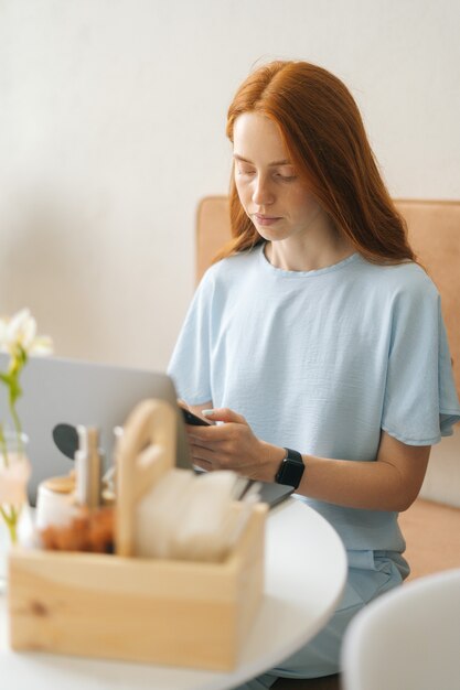 Vertical portrait of focused young woman in smart watch using laptop and phone sitting at table in cafe with warm daylight, selective focus. pretty redhead lady remote working or studying indoors.