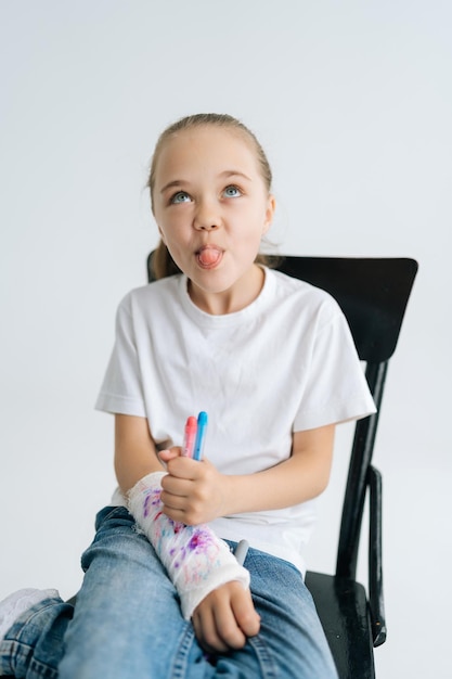 Vertical portrait of fooling little girl drawing cute image with colorful marker on broken hand wrapped in white plaster bandage and showing tongue