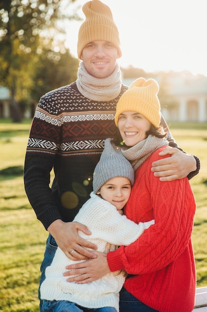 Vertical portrait of friendly family couple stand together embrace each other have good relationships enjoy sunny weather Handsome unshaven man hugs his wife and daughter pose outdoors