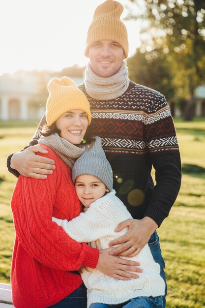 Vertical portrait of friendly family couple stand together embrace each other have good relationships enjoy sunny weather Handsome unshaven man hugs his wife and daughter pose outdoors