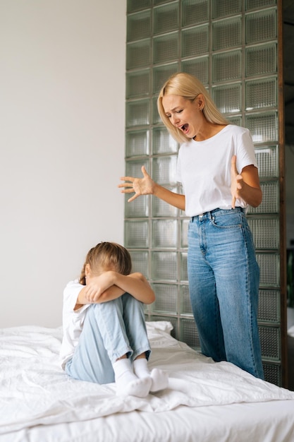 Vertical portrait of furious young mother scolding screaming for bad behavior to depressed crying little girl covering face with palm sitting on bed at home