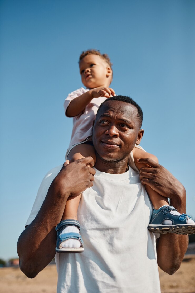 Vertical portrait of happy africanamerican father carrying son on shoulders against blue sky while e...