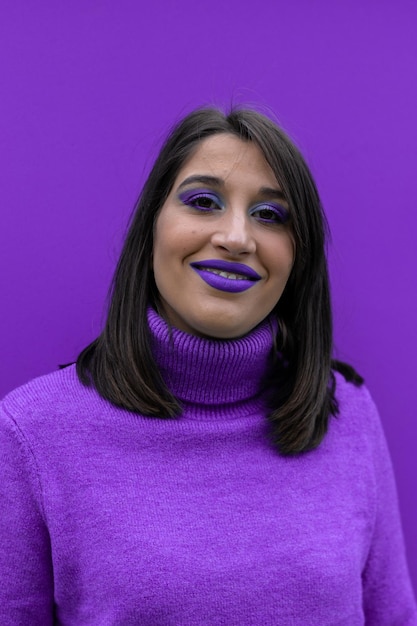 Вертикальный портрет счастливой молодой женщины, улыбающейся и смотрящей в камеру, одетой и накрашенной в фиолетовый цвет на фиолетовом фоне