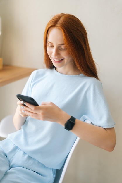 Vertical portrait of laughing young woman wearing casual clothing using mobile phone sitting at desk by window in cozy light cafe. Pretty redhead Caucasian lady having leisure activity in coffee shop.