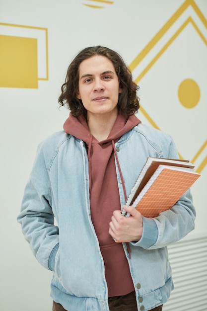 Vertical portrait of long haired young man in school looking at camera while standing against wall and holding notebooks