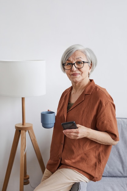 Ritratto verticale di una moderna donna d'affari senior che guarda la telecamera e si gode il caffè in interni minimi