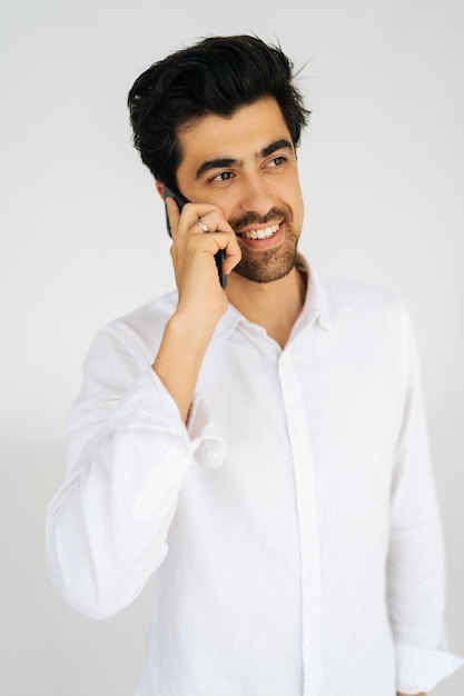 Vertical portrait of positive young man in shirt talking mobile phone having conversation on smartphone smiling looking away