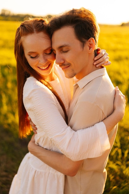 Vertical portrait of romantic young couple in love hugging kissing standing together on green meadow in summer evening during golden sunset