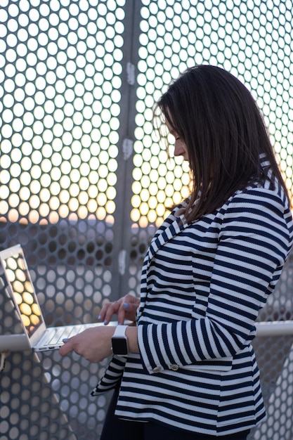 日没時に彼女のコンピューターで外で働く笑顔の白人女性の縦の肖像画