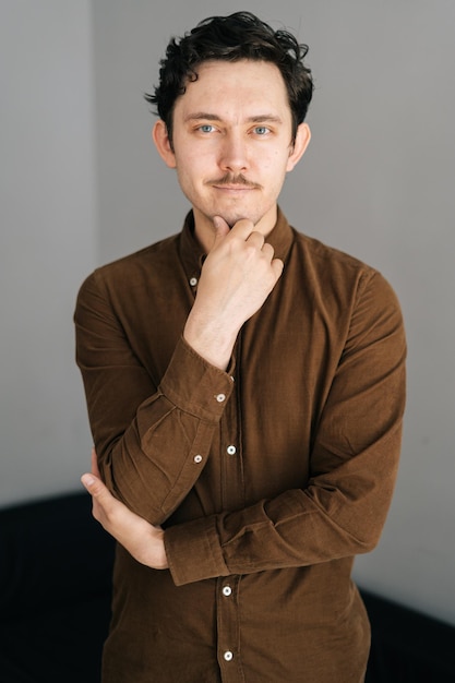 Vertical portrait of smiling young man with little moustache holding hand on chin and thoughtful looking at camera thinking over solution