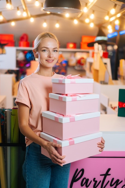 Vertical portrait of smiling young woman holding many beautiful Christmas gift boxes on background of holiday shop interior looking at camera