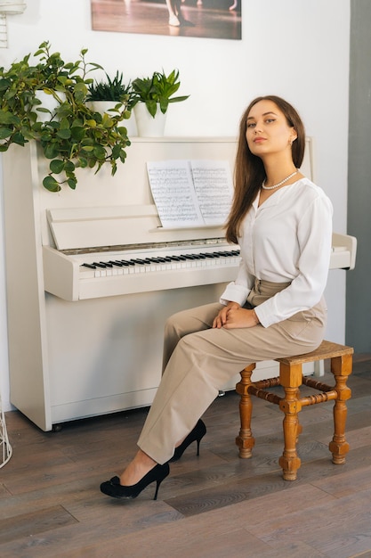 Vertical portrait of successful young woman pianist looking at camera sitting at classical white piano in classroom Satisfied attractive female musician performer preparing for classes at home studio