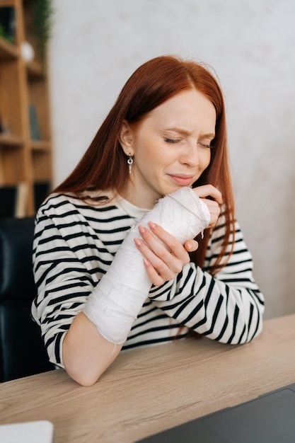 Vertical portrait of suffering from pain crying young woman with broken right hand wrapped in gypsum bandage sitting at table closed eyes