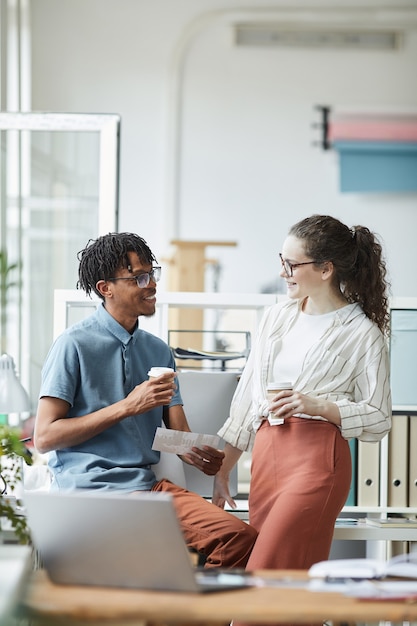 Photo vertical portrait of two creative young people looking at printed photographs and chatting during coffee break in modern office