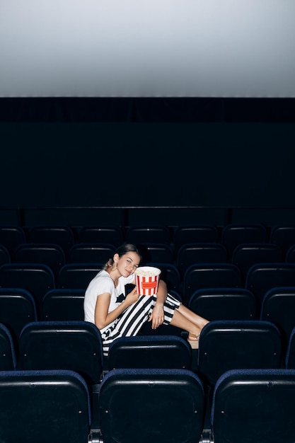 Foto ritratto verticale di una donna seduta su una sedia in un cinema che mangia una ciotola di popcorn