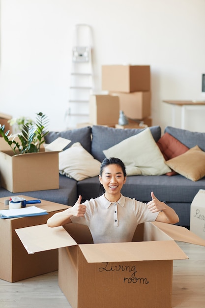 Vertical portrait of young Asian woman sitting in box and showing thumbs up while moving in to new house or apartment