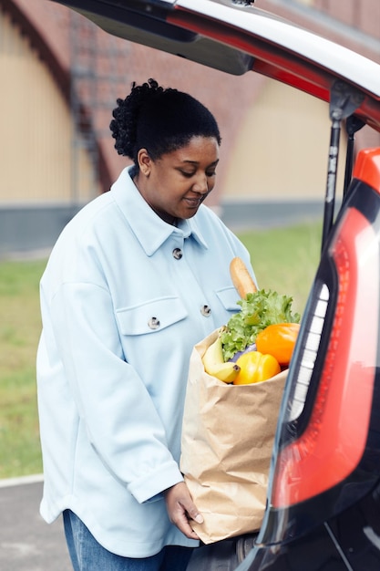 Ritratto verticale di giovane donna nera che mette la borsa della spesa nel bagagliaio dell'auto al parcheggio del supermercato