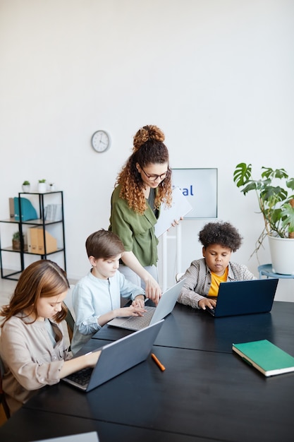 Ritratto verticale di una giovane insegnante che aiuta i bambini a utilizzare i laptop durante le lezioni di informatica a scuola