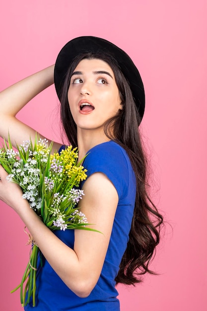 Vertical portrait of young girl wearing hat and holding bunch of flowers on pink background