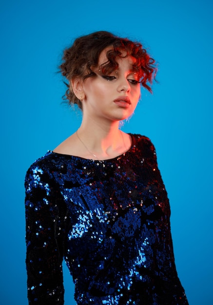 Vertical portrait of young lady standing on blue background and looking aside curlyhaired girl in gilded dress high quality photo