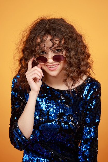 Vertical portrait of a young lady wearing gilded dress and sunglasses on orange background High quality photo