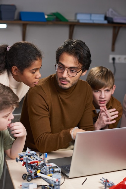 Ritratto verticale di giovane insegnante maschio in classe di robotica con diversi gruppi di bambini intorno