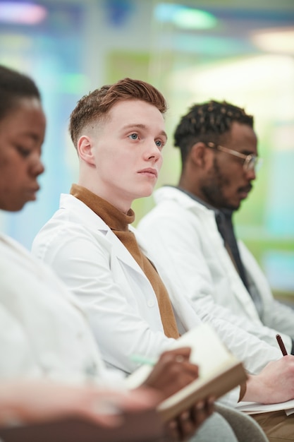 Vertical portrait of young man wearing lab coat while sitting in row in audience and listening to lecture on medicine in college